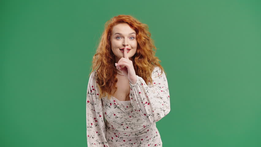 Beautiful woman with natural red curls makes shush shh gesture with finger on lips, silence signaling