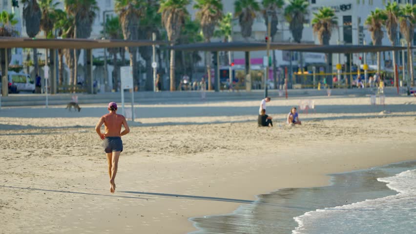Middle age man running or jogging on sandy beach in early morning in Tel Aviv. Active healthy person training outdoors on summer day in Israel by Mediterranean Sea. Palm trees and hotels in background