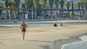 Middle age man running or jogging on sandy beach in early morning in Tel Aviv. Active healthy person training outdoors on summer day in Israel by Mediterranean Sea. Palm trees and hotels in background - Powered by Shutterstock - Get 15% off with code: PIKWIZARD15