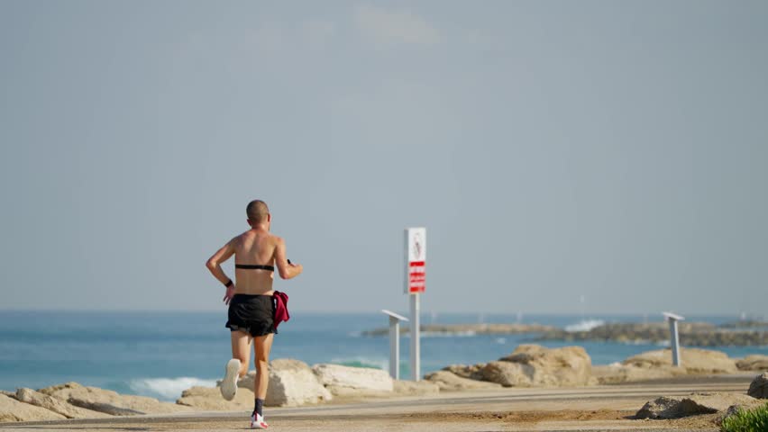 Person exercizing jogging and running on beach promenade in Tel Aviv Israel. Man training on hot summer day outdoors by the Mediterranean Sea. Determined and serious in early morning sun by blue sea