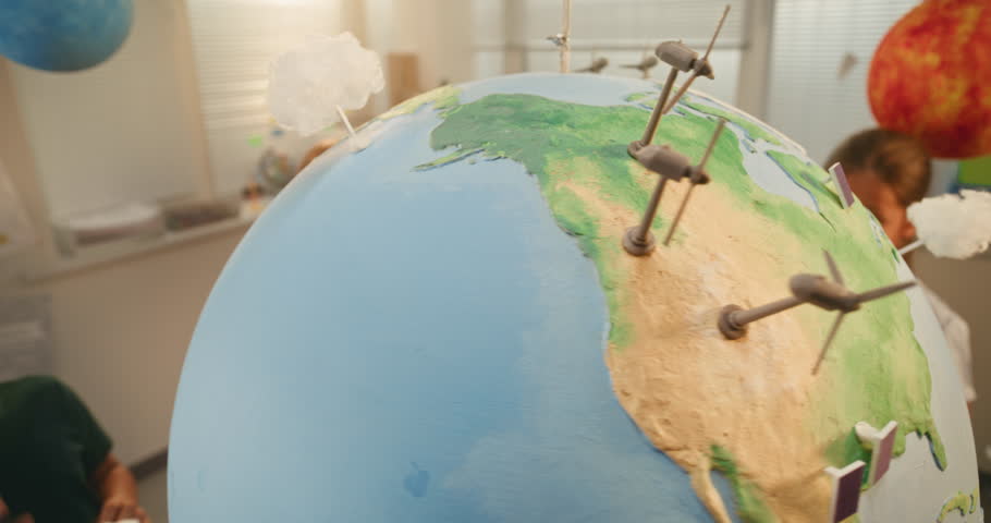 Renewable Energy STEM Class: Close Up of Earth Model with 3D Models of Wind Turbines and Solar Panels in Modern Classroom. Elementary School Students Attaching Miniature Handmade Clouds to Globe.