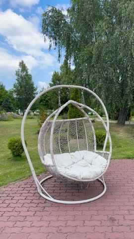 A comfortable wicker swing chair with a soft cushion stands on a patio in a beautiful summer park, inviting for relaxation.Modern garden furniture: a cozy hanging egg chair for two