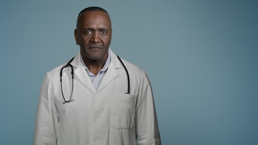 African American man doctor male physician pointing finger at empty space for writing text standing in blue studio template for advertising medical services healthcare promotion professional treatment