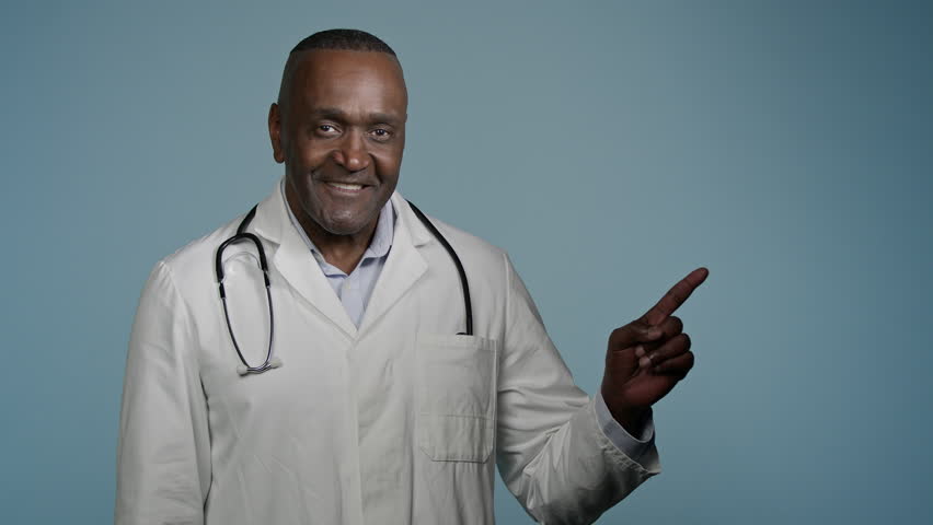 African American man doctor male physician pointing finger at empty space for writing text standing in blue studio template for advertising medical services healthcare promotion professional treatment