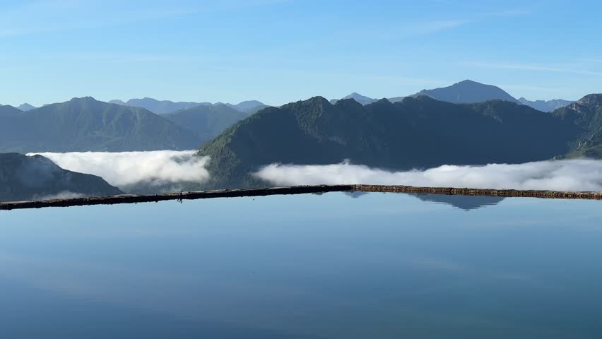 Calm waters mirroring majestic mountain range and soft cloud layers through pristine infinity pool during luminous summer morning
