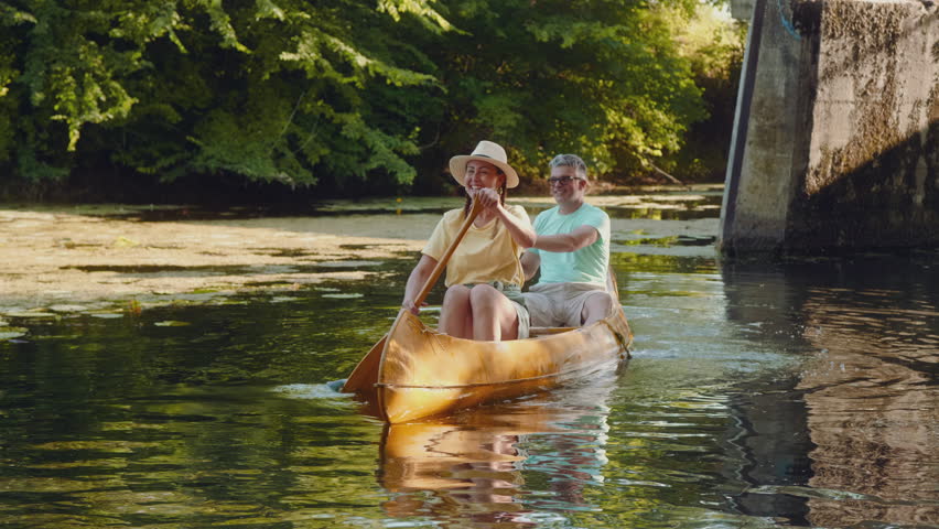 Excited middle-aged woman raises her oar, looking forward to the achievement of canoeing with a friend on a river.