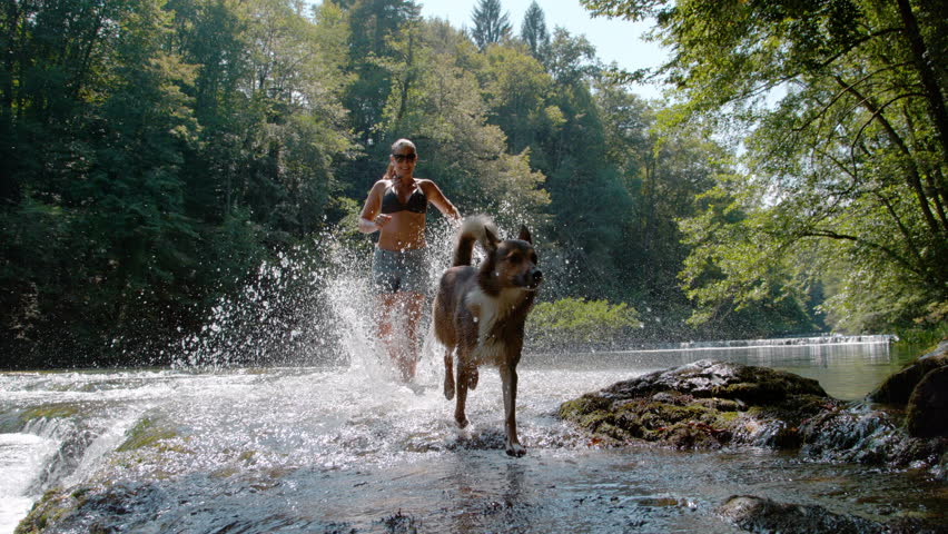 SLOW MOTION, PORTRAIT: Smiling woman and shepherd dog run through shallow river water and create big splashes of flying drops. Refreshing outdoor activity in the wilderness on a hot, sunny summer day.