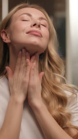 Happy young woman girl applying cream on face skincare look mirror at bathroom Caucasian female model customer touching soft skin antiaging lotion serum massaging neck hands beauty routine cosmetics