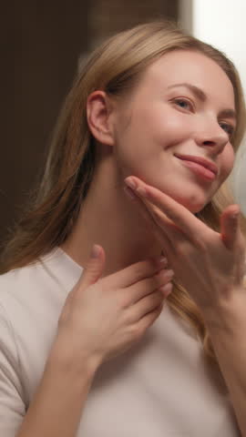 Happy young woman girl applying cream on face skincare look mirror at bathroom Caucasian female model customer touching soft skin antiaging lotion serum massaging neck hands beauty routine cosmetics