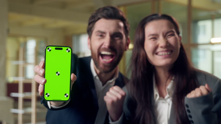 Excited two business woman man celebrating win showing mobile phone green screen chroma key mock up Caucasian Korean businesspeople cheering winner victory money smartphone excitement triumph office