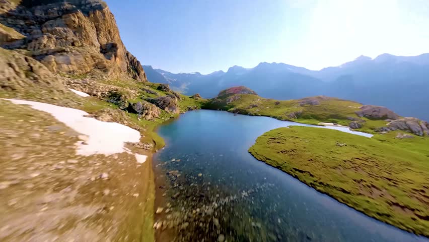 Cinematic FPV drone footage flying over a green grassy hill with rocky mountains under clear blue sky, capturing stunning outdoor nature landscape scenery. Perfect for travel videos, nature documentar