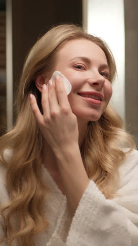 Happy beautiful Caucasian woman wiping skin face cotton pad toner skincare smiling girl female model bathrobe cleansing makeup sunscreen removal lotion micellar water acne treatment at home bathroom