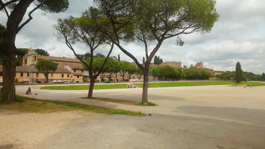 View of the ancient Circus Maximus with trees and ruins in the background under a partly cloudy sky.