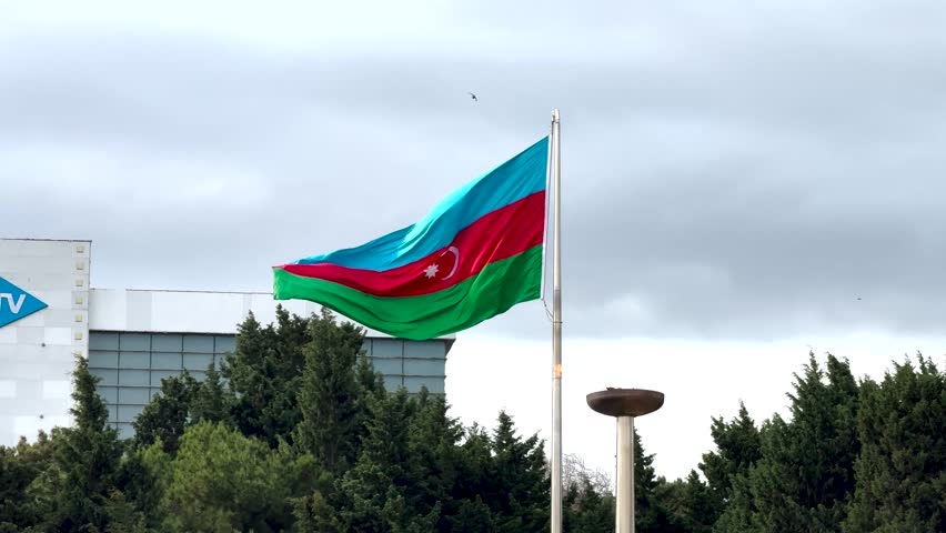 National flag of Azerbaijan fluttering beside the state TV building and pine trees. Baku, Azerbaijan