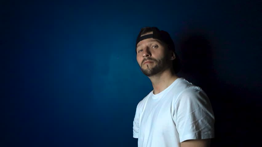 Man with white shirt black reversed cap hat and tattoos holds up a CPL polarizer filter and looks through it with confidence and happiness in slow motion while being in front of a blue studio wall.