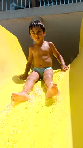 Energetic young boy sliding down vibrant yellow waterslide, splashing joyfully into cool blue pool during sunny summer day at water park