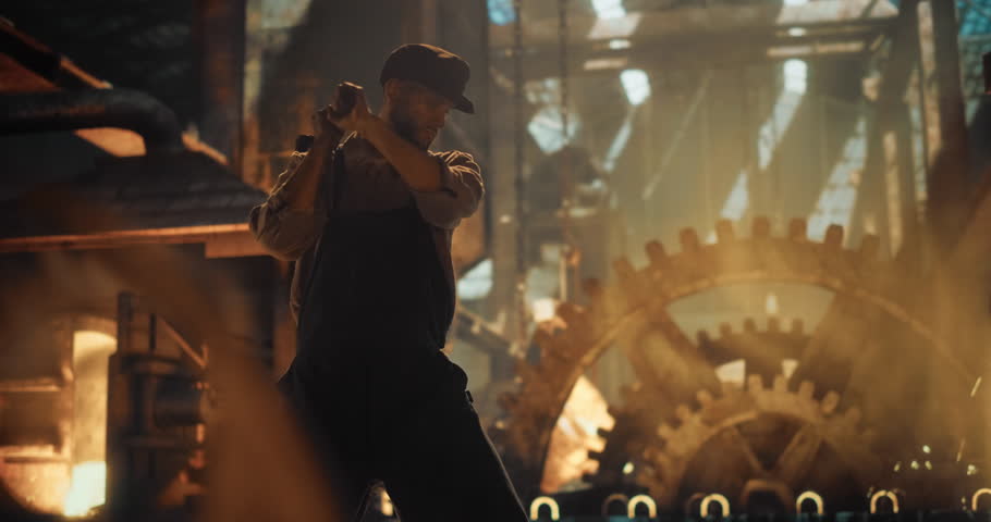 Man in Worn Work Clothes Strikes Iron with a Sledgehammer Inside a Gritty, Fire-Lit Workshop. Man s Doing Manual Labor During the Industrial Revolution. Cinematic Slow Motion Footage