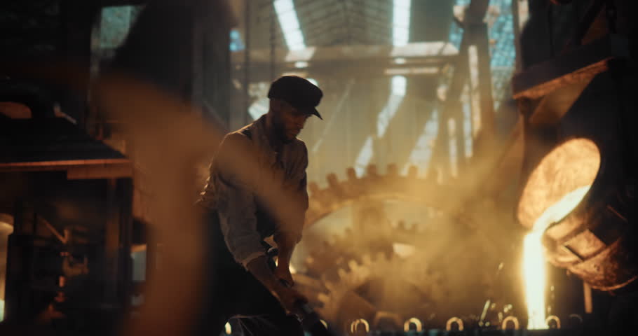 Blacksmith or Foundry Worker from the 1800s Uses a Sledgehammer to Shape Metal, Drive Wedges or Breaking a Fresh Mold. Dark, Smoky Atmosphere Lit by Furnace Fire. Slow Motion Footage