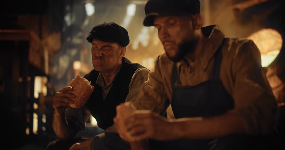 Worn-Out Male Factory Workers of the Industrial Revolution Pause For a Lunch Break to Eat a Loaf of Bread by the Furnace. Their Clothing is heavily Covered in Soot, Expressions on Faces Shows Fatigue