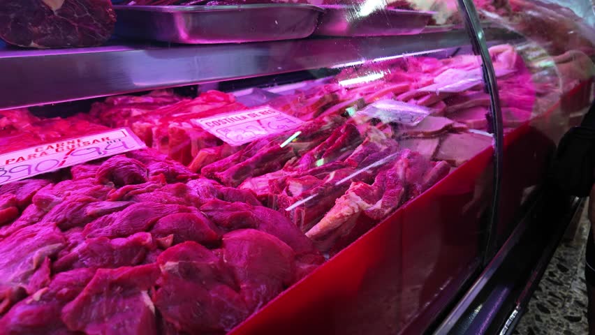 refrigerated display case at butcher shop filled with various cuts of fresh red meat under pink lighting.
