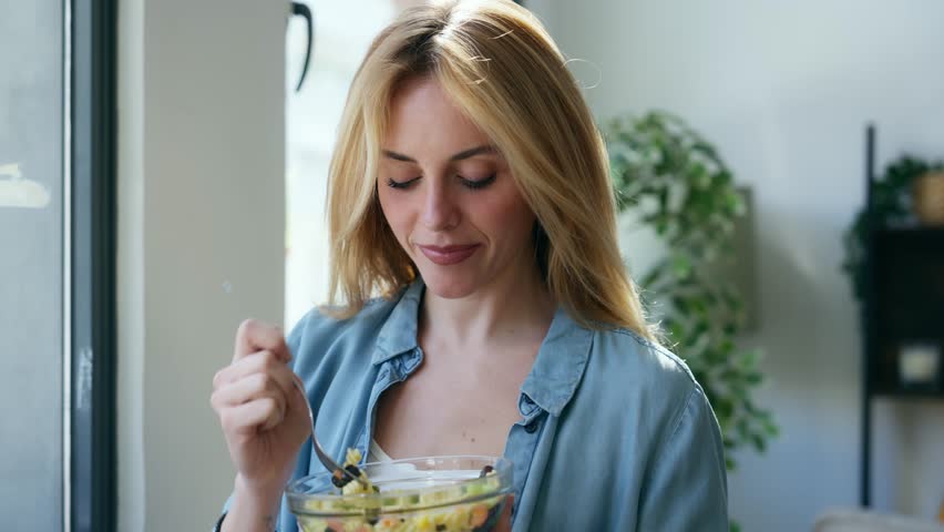 Video of smiling woman eating healthy pasta salad while sitting on the living room at home