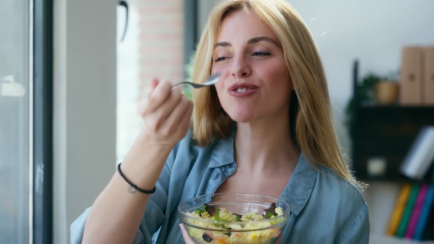 Video of smiling woman eating healthy pasta salad while sitting on the living room at home