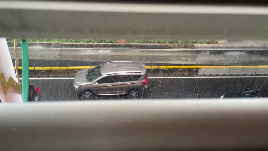 Jakarta, June 16, 2025. A rainy street scene viewed through window blinds, with cars driving below and a pedestrian bridge visible in the background during a downpour.