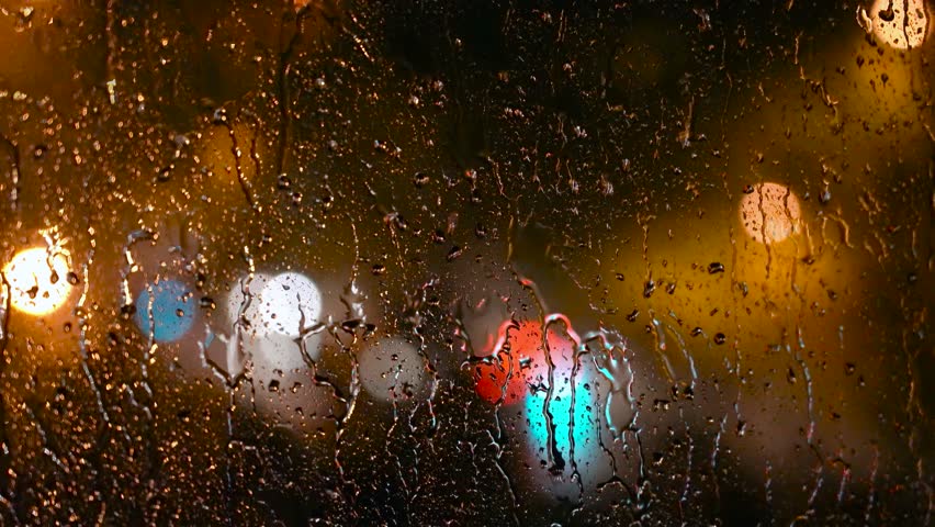 City Lights at Rainy Night Seen Through a Window with Rain Drops - Rack Focus. Rainstorm in the City at Night. Blurred City Night Illumination Through the Home Window with Raindrops. Melancholy Rain.