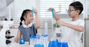 Asian girl watching as Asian boy lifts test tube with blue liquid during slowmotion science experiment inside bright elementary classroom filled with lab tools and collaborative learning - Powered by Shutterstock - Get 15% off with code: PIKWIZARD15