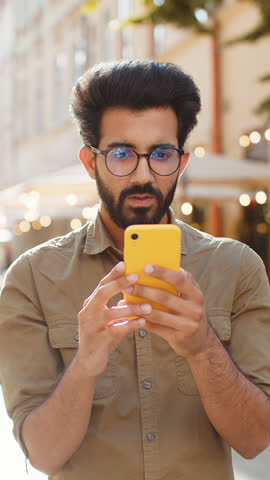 Indian bearded man use smartphone typing browsing, loses becoming surprised sudden lottery results, bad news, fortune loss, fail. Guy walking in urban city street outdoors. Town lifestyles. Vertical