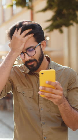 Indian bearded man use smartphone typing browsing, loses becoming surprised sudden lottery results, bad news, fortune loss, fail. Guy walking in urban city street outdoors. Town lifestyles. Vertical