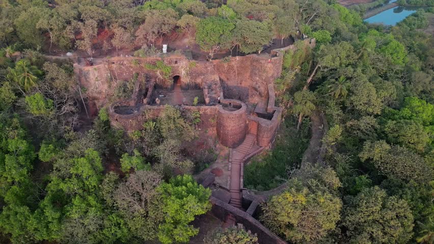 Remains of Chapora Fort nestled in Goa's dense forest. Colonial-era fort entrance with round towers above forested slope. Calm waters shape curved sandbanks at Goa river mouth.