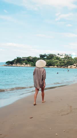 Relaxed woman tourist walking alone on sandy beach in morning, vertical video, back view. Traveling and exploring new places, memories from summer vacation, female holidaymaker relaxing on shore alone