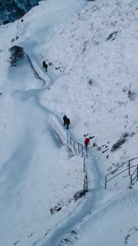 A vertical video taken from above of a group of mountaineers as they climb to base camp on the Mardi Himal trek in Nepal. The mountains are covered in thick snow, and footprints form a path.