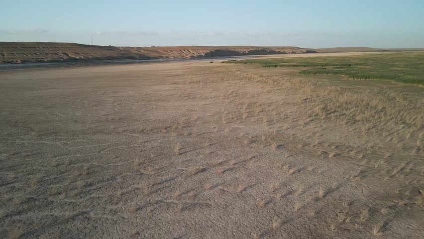 Aerial view of over parched land dotted with dry grass. In the distance, green vegetation forms a striking border. The arid landscape meets life-giving water of Amu Darya in Uzbekistan's desert region