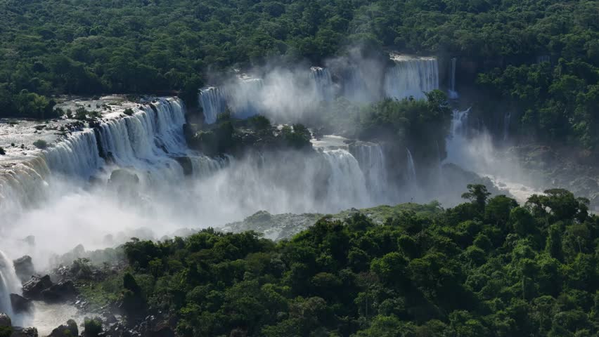 Aerial view reveals Iguazu Falls in Argentina. Water pours over cliffs into dense jungle. Mist floats above as the river splits into many falls forming a natural spectacle