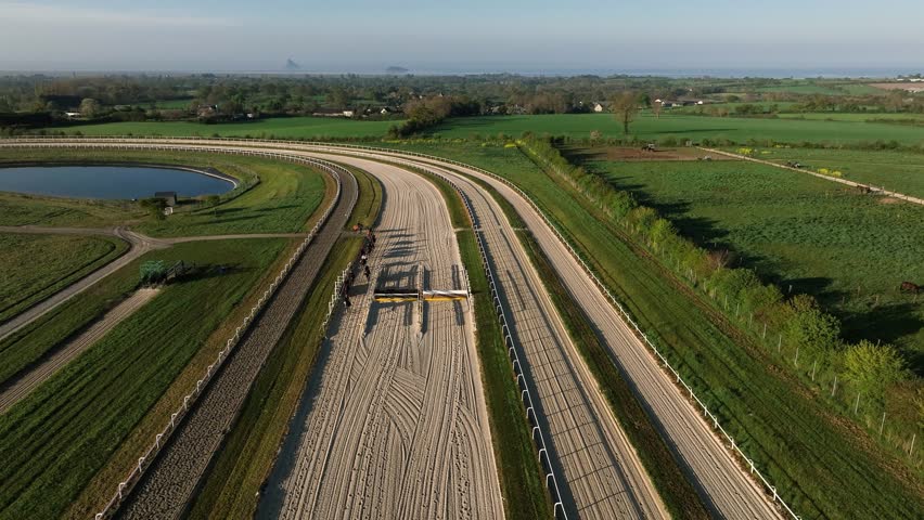 drone footage of horses and riders galloping on a racetrack in the cool air of an early Normandy morning.