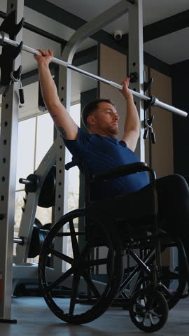 Focused man in wheelchair performs overhead barbell press in gym