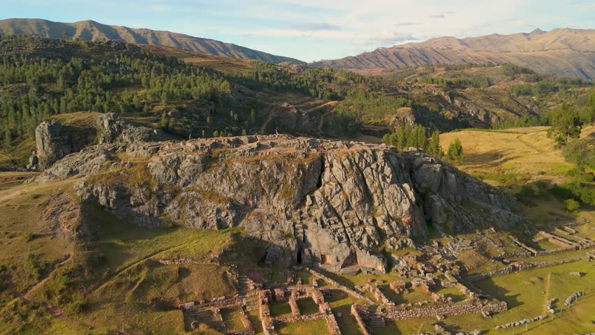 Aerial parallax shot of the Temple of the Moon near Cusco, Peru, with tourists visible in the distance exploring the ancient Inca ruins and dramatic rocky landscape.