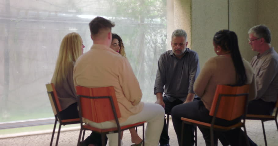 Diverse group of adults sit in circle during emotional group therapy session, engaging in serious conversation in quiet, supportive indoor setting with natural light