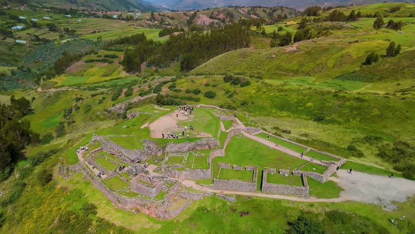 An aerial parallax drone shot circles the Puka Pukara archaeological site, revealing tourists exploring the ancient stone walls and terraces of the historic Inca 'Red Fortress' near Cusco, Peru.