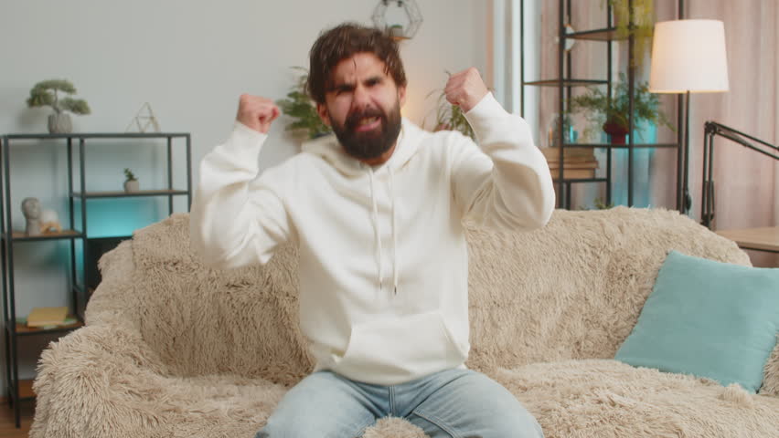 Happy Indian young man watching TV shouting, celebrating success, winning, goal achievement good news, lottery luck, victory sitting on sofa in living room. Adult guy clenching fists in home room.