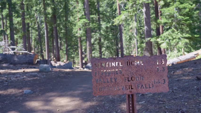 A scenic view of a metal hiking trail signpost in Yosemite National Park, California, showing distances to Sentinel Dome, Fissures, Bridalveil Falls, and the valley floor
