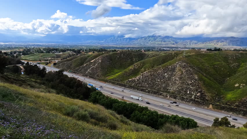 Timelapse of rolling green hills beside a busy highway as vehicles travel in the valley below. A picturesque sky features scattered clouds, adding depth to the landscape.
