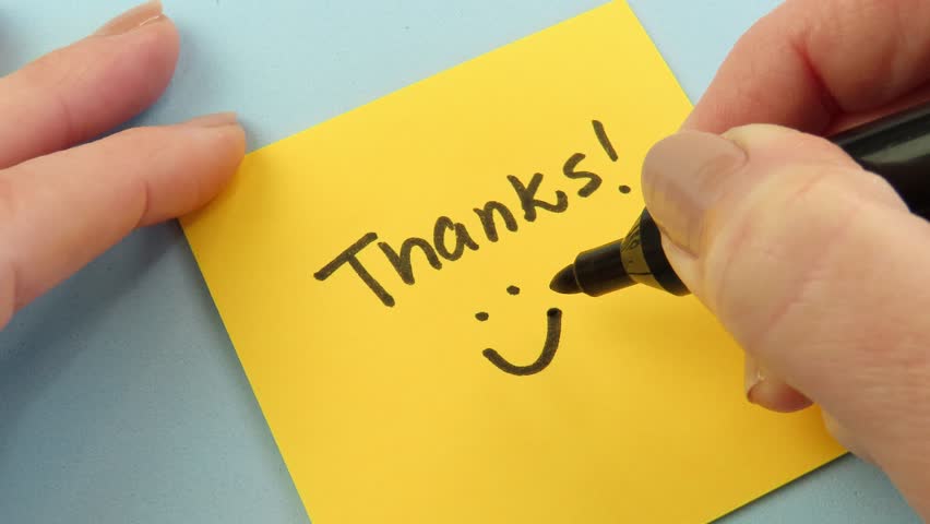 Video of woman writing the expression "Thanks" and drawing a smile on a sticky note. Thank you note.