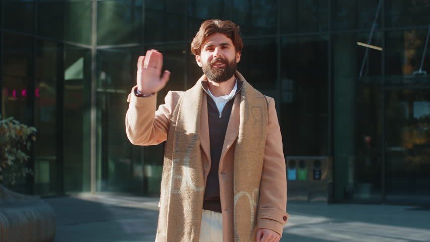 Cheerful Arabian man tourist smiling friendly at camera, waving hands gesturing invitation hello hi, greeting, goodbye, welcoming with hospitable expression outdoors. Happy guy on downtown city street