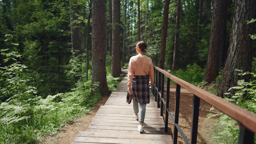 Visit natural park in summer, woman tourist walking on wooden path in forest. Female traveler exploring unique natural location in reserve park area, rest in eco glamping in woodland, back view