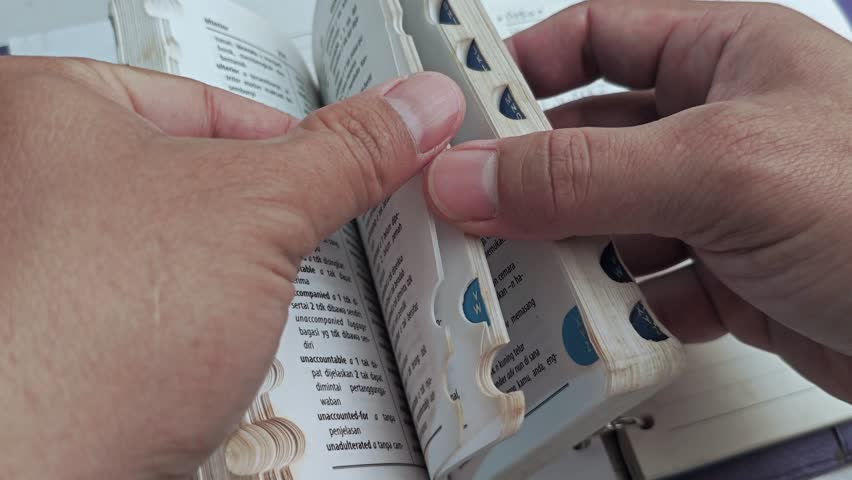 Close-up of hands flipping through a dictionary, searching for a word's meaning. Focus on pages, text, and the action of looking up definitions.