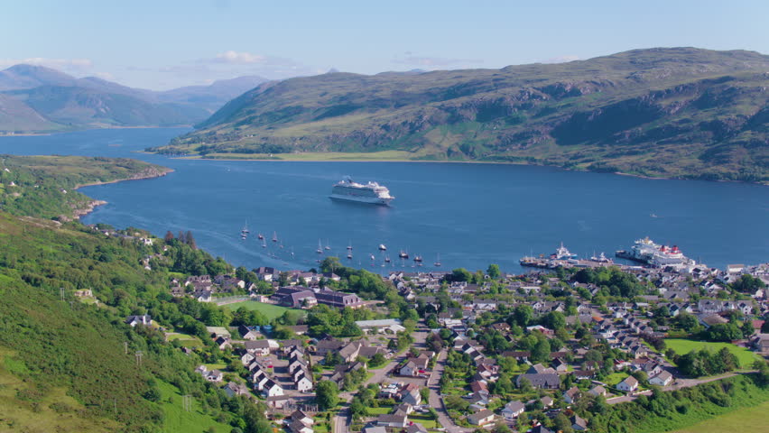 Drone shot of Viking Sun cruise ship anchored in the loch near Ullapool, with stunning views of the village, surrounding mountains, and clear blue skies on a beautiful day.