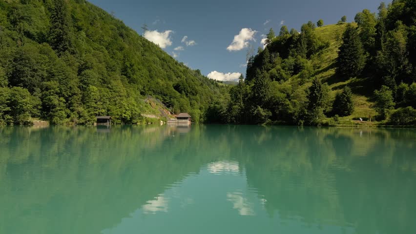 A low drone view of Klammsee Lake that reflects the surrounding green, forested mountains and a clear, blue sky with scattered clouds in Austria